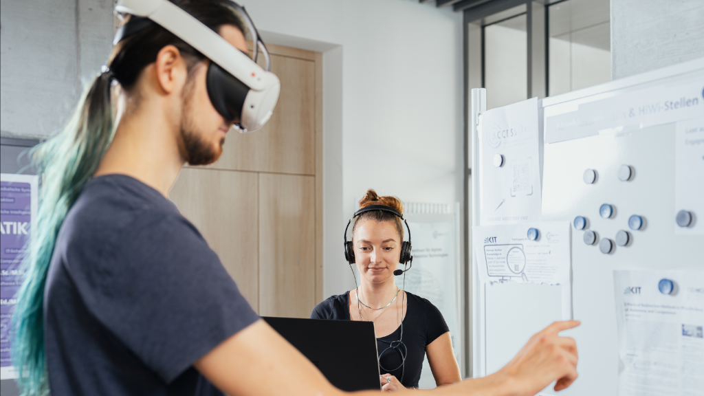 Person mit VR Brille zeigt auf Aushängetafel. Im Hintergrund steht eine Person mit Headset und schaut auf ihren Laptop.